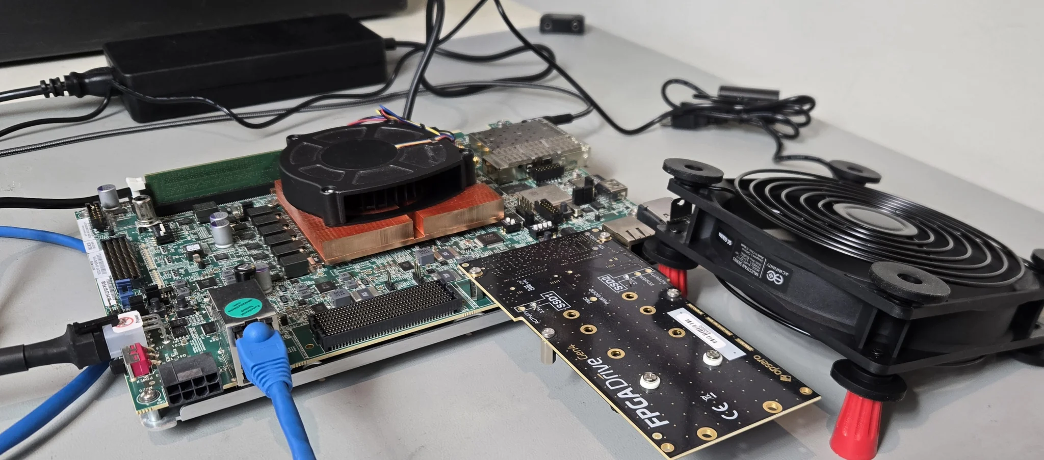 The AMD Versal AI Core VCK190 evaluation board with the Opsero FPGA Drive FMC Gen4 (OP063) in its FMCP1 connector; the two Sandisk WD_BLACK SN8100 NVMe SSDs are mounted on the underside of the card (not visible), and a desk fan beside the board blows air over them.