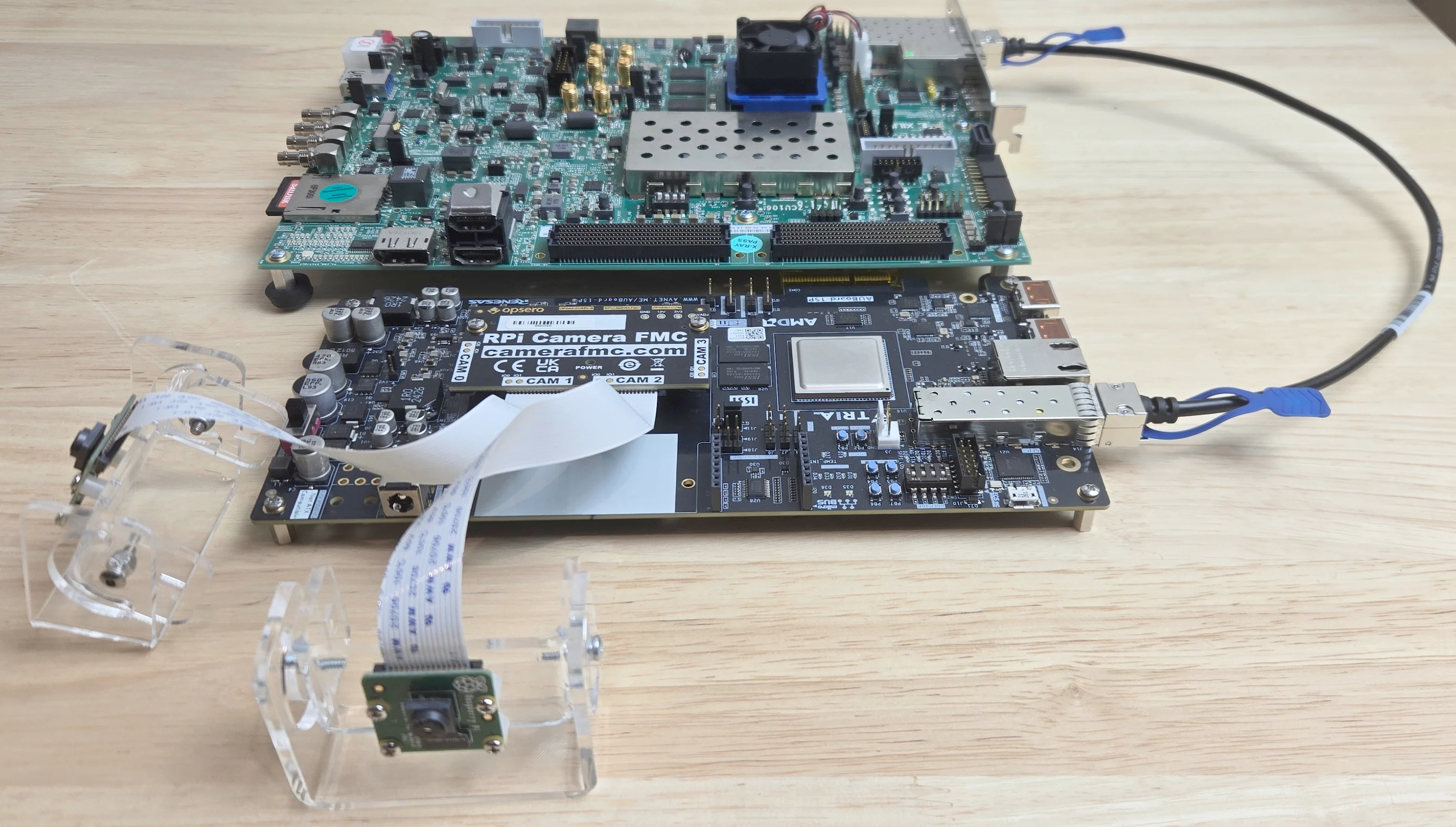 Photo of the proof-of-concept setup: an AMD ZCU106 and a Tria AUBoard joined by an SFP+ DAC cable, with the RPi Camera FMC on the AUBoard and two Raspberry Pi cameras on acrylic stands